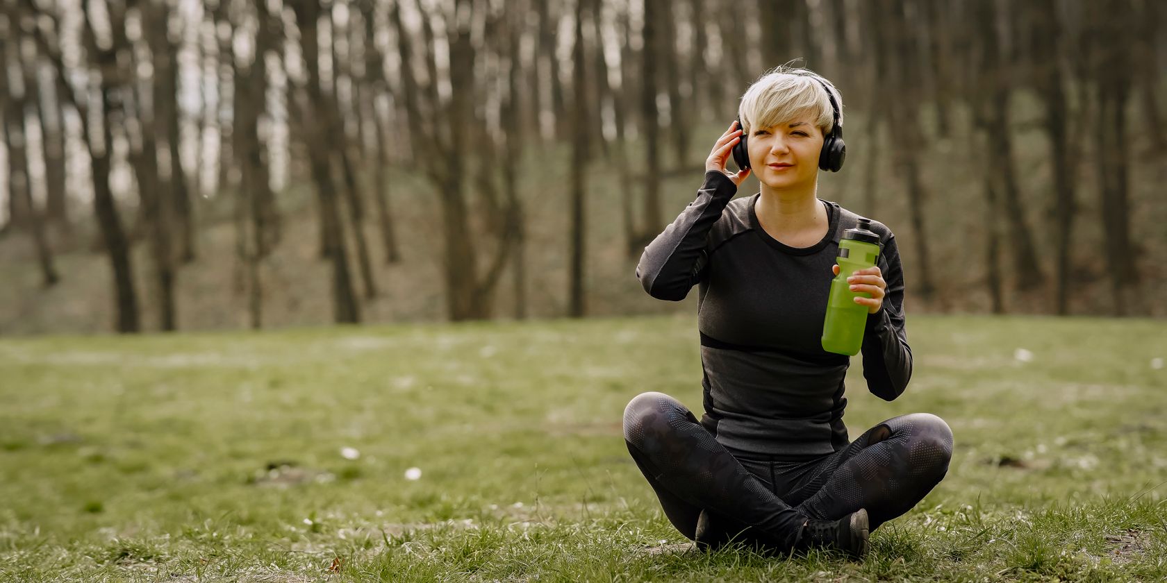 Uma mulher sentada ao ar livre na natureza bebendo água e ouvindo fones de ouvido